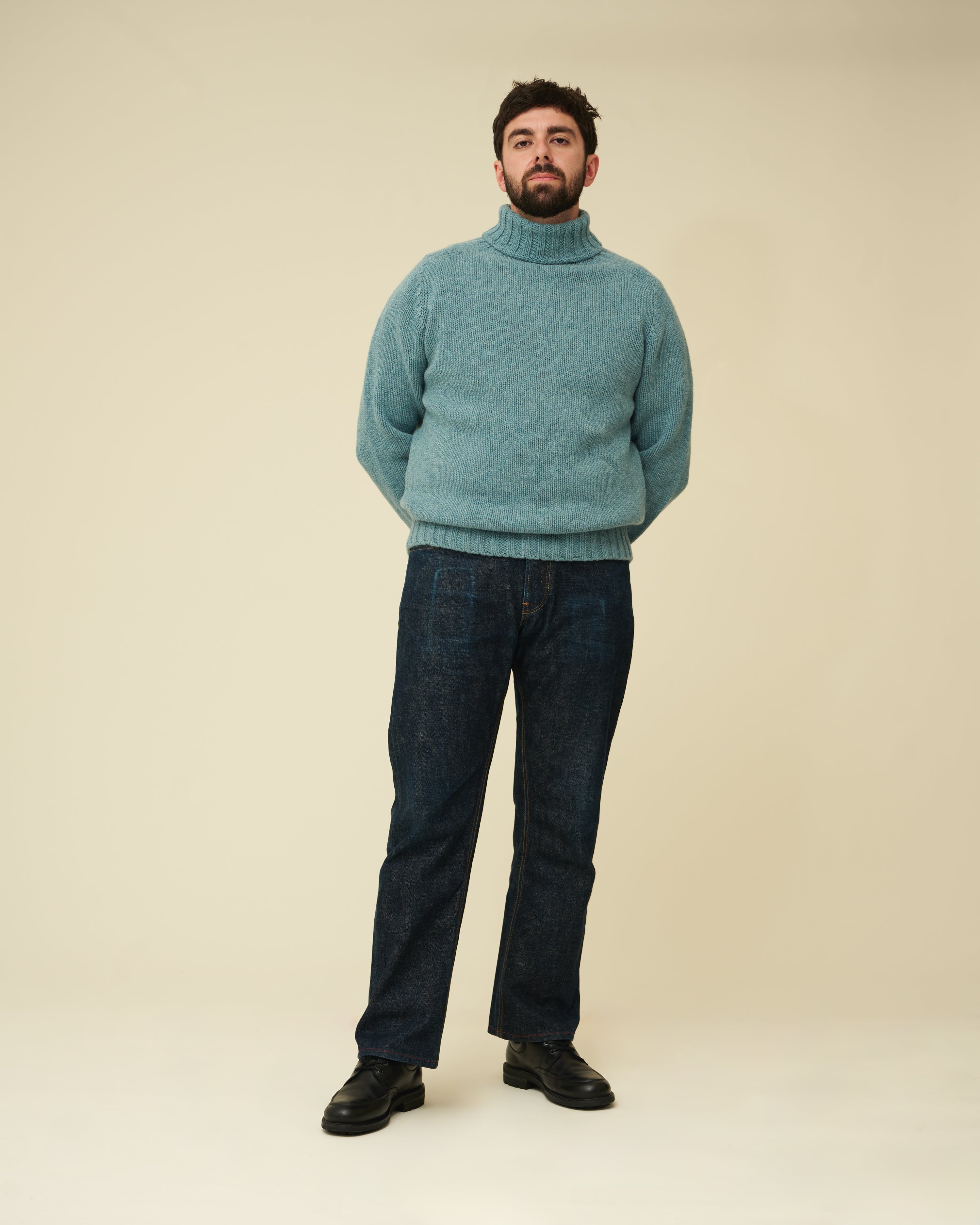 Man wearing a teal sweater and dark jeans on a beige background
